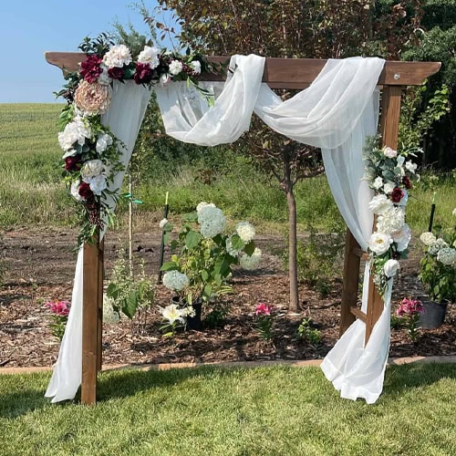 Beautiful wooden wedding arch decorated with white and burgundy flowers, flowing white fabric for outdoor ceremonies.